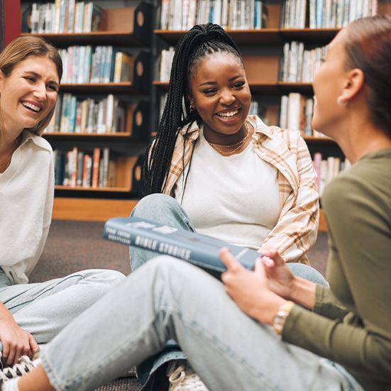 Tre unge kvinder i et bibliotek sidder på gulvet og taler sammen