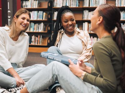 Tre unge kvinder i et bibliotek sidder på gulvet og taler sammen